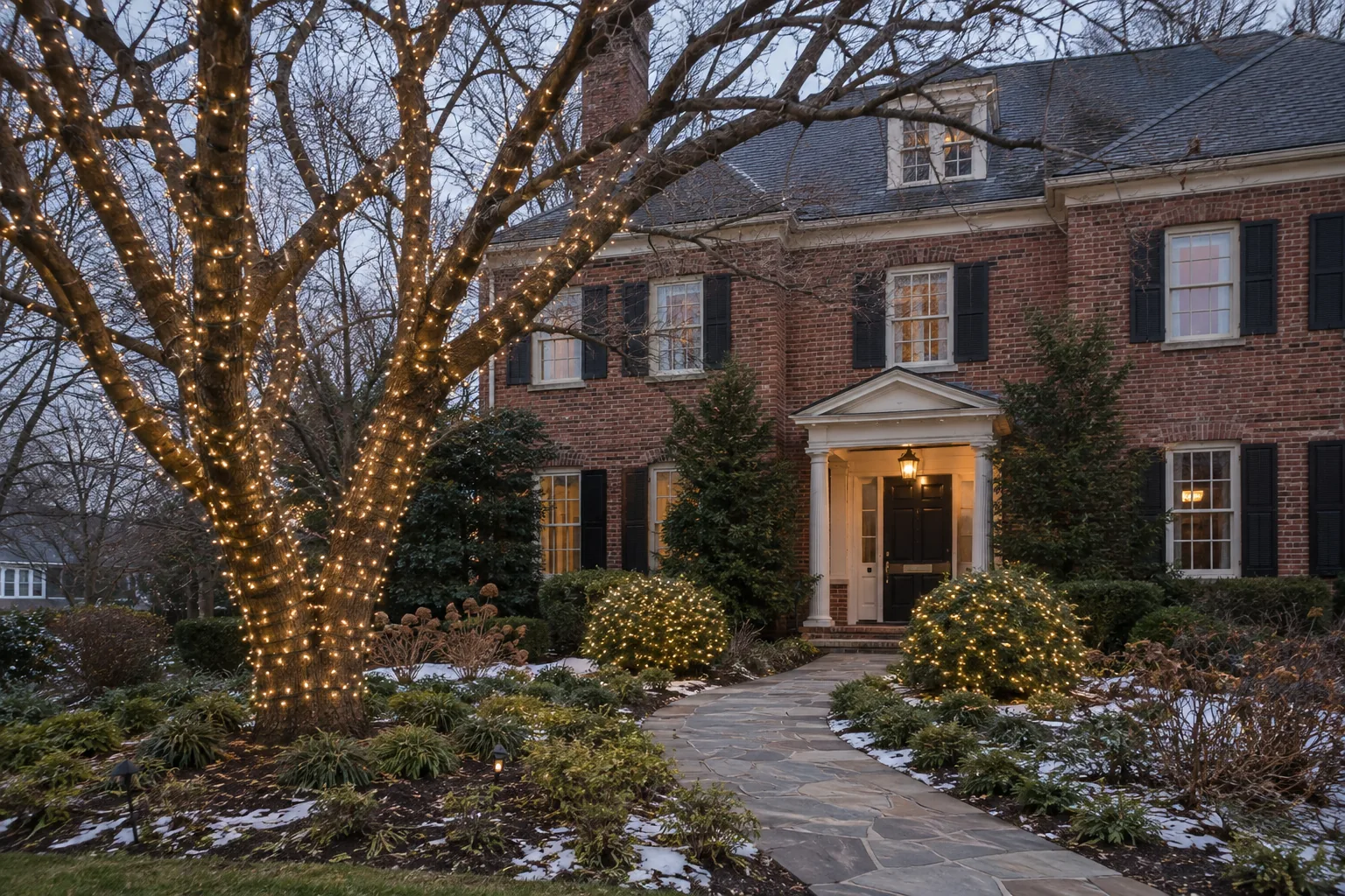 mature tree and shrubs wrapped in small warm white lights beside a brick home
